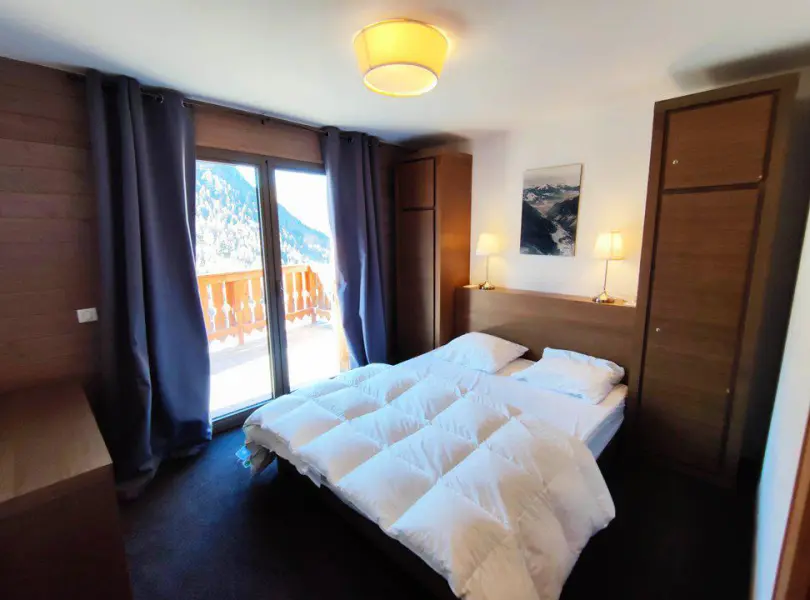Chambre cosy avec lit double, armoire en bois, lampes de chevet et accès à un balcon avec vue sur la montagne.