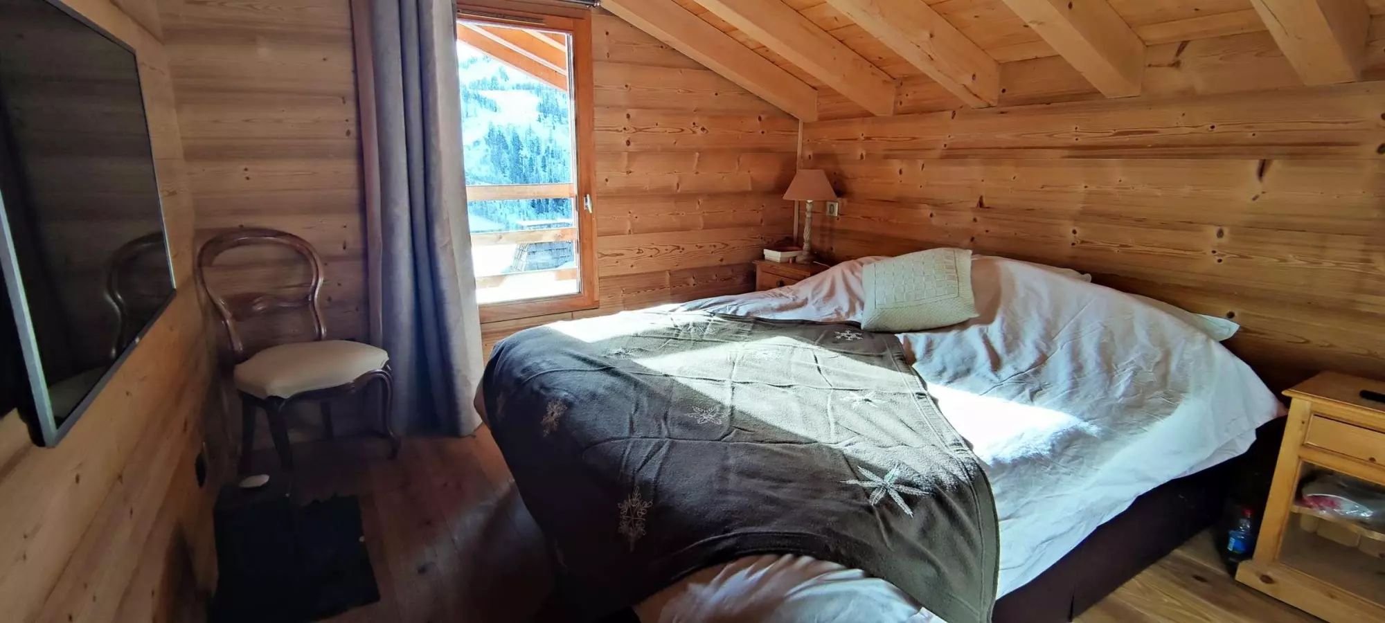 Chambre en bois avec lit double, couverture à motifs et fenêtre offrant une vue sur les montagnes enneigées.