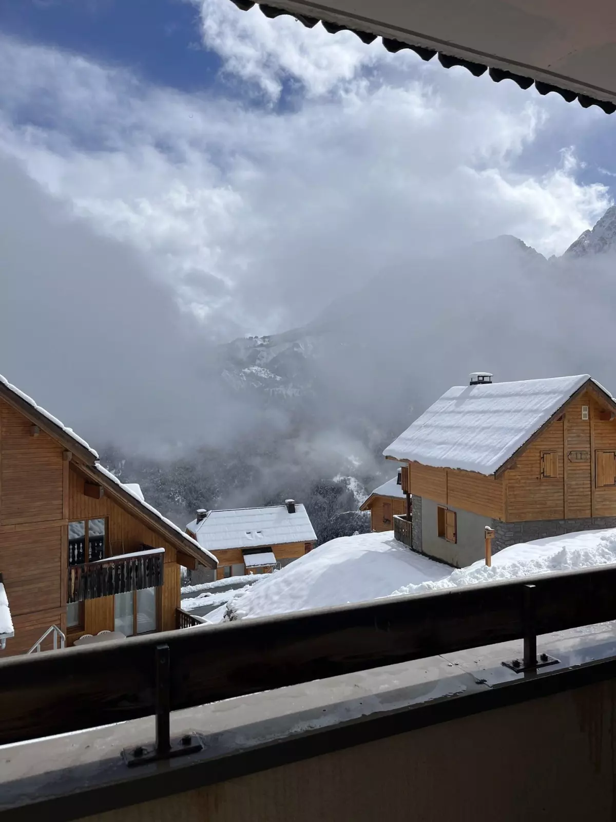 Vue depuis un balcon sur un village de chalets en bois enneigés, entouré de montagnes et de nuages dans une station alpine.