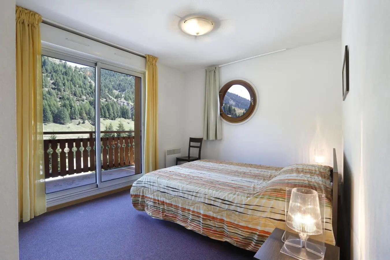 Chambre avec lit double, grande baie vitrée ouvrant sur un balcon en bois et vue imprenable sur la montagne verdoyante.