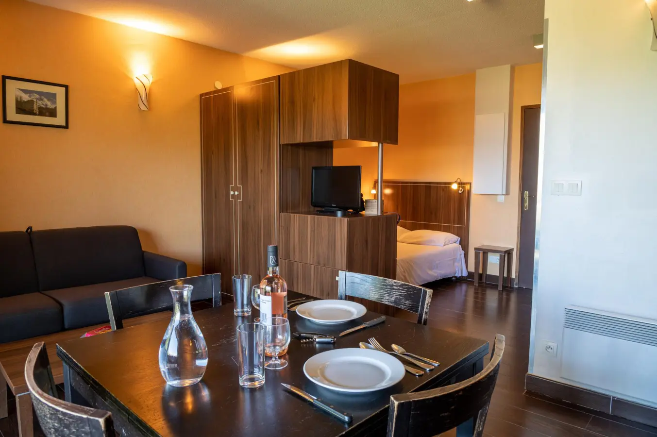 Appartement moderne avec coin repas dressé, salon avec canapé et télévision, et espace chambre séparé par une armoire en bois