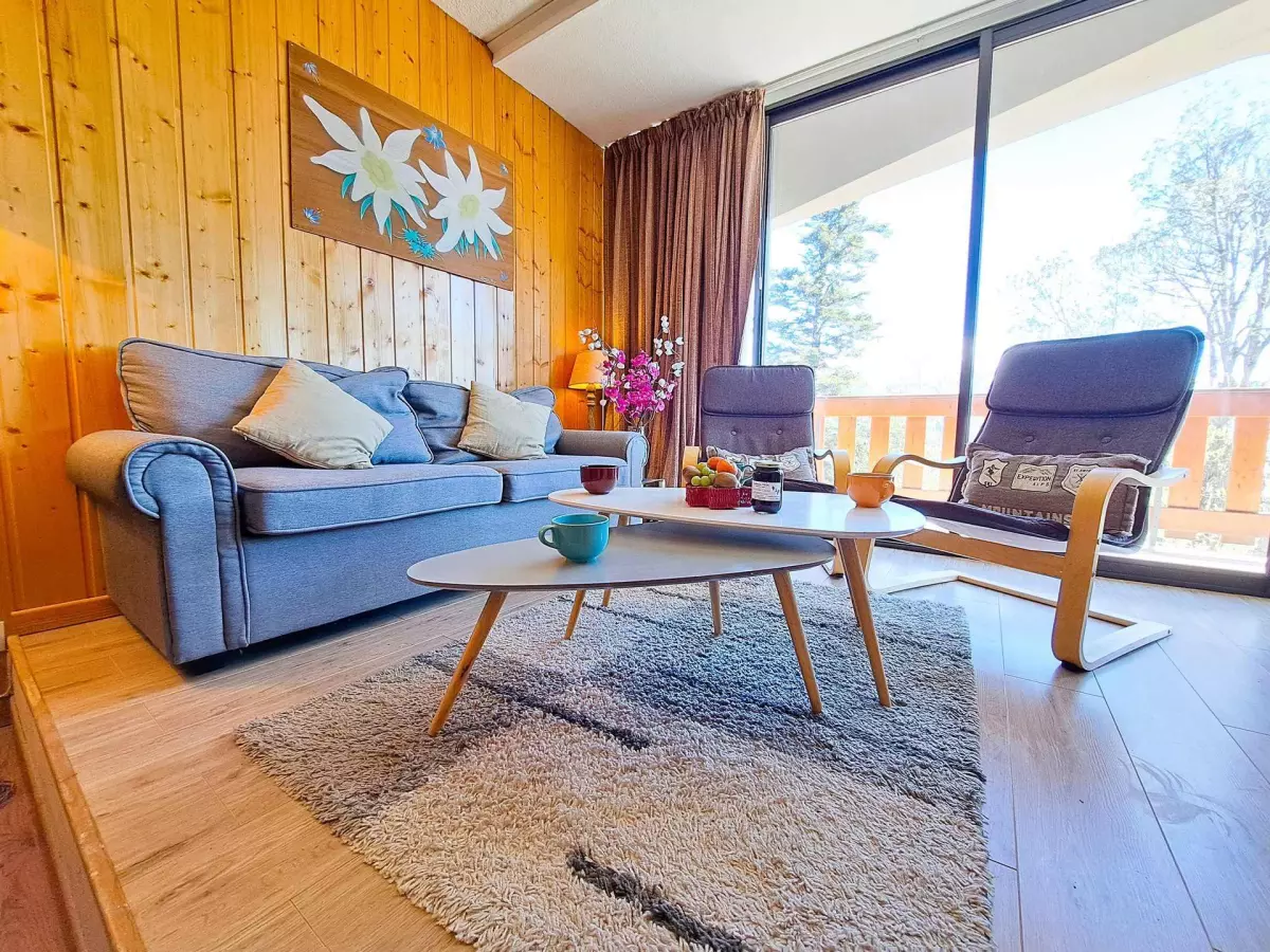 Salon lumineux avec canapé confortable, fauteuils modernes et grande baie vitrée donnant sur un balcon en bois avec vue sur la nature.