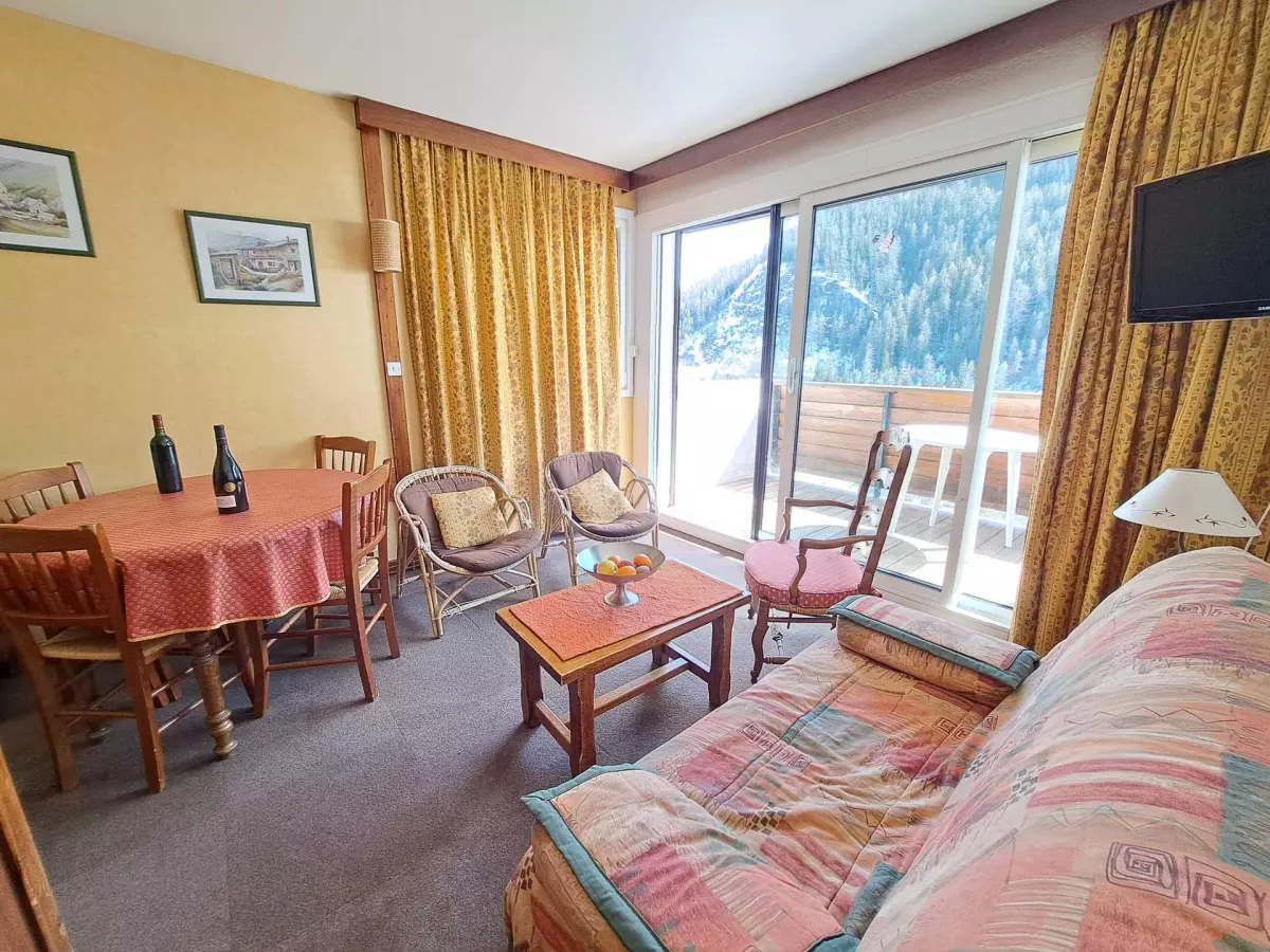 Salon de montagne au style vintage avec coin repas, canapé coloré et accès à un balcon ensoleillé offrant une vue sur la forêt enneigée