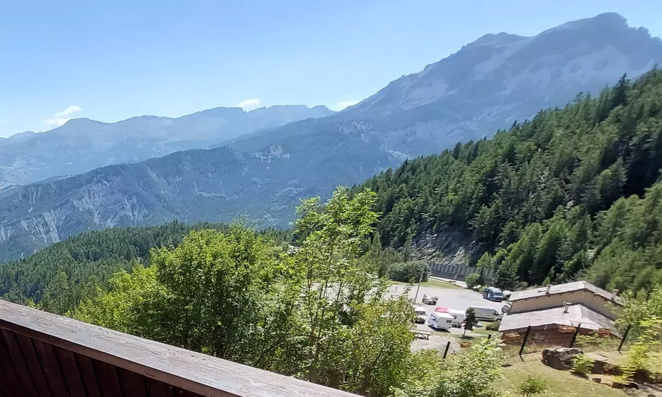 Vue panoramique depuis un balcon sur les montagnes et forêts alpines, avec un parking et quelques chalets en contrebas sous un ciel dégagé