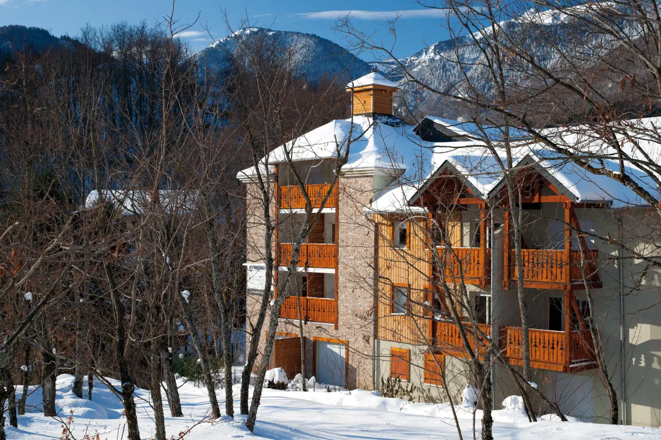 Résidence Lagrange Les Chalets d'Ax en hiver, entourée d'arbres et de montagnes enneigées avec balcons en bois.