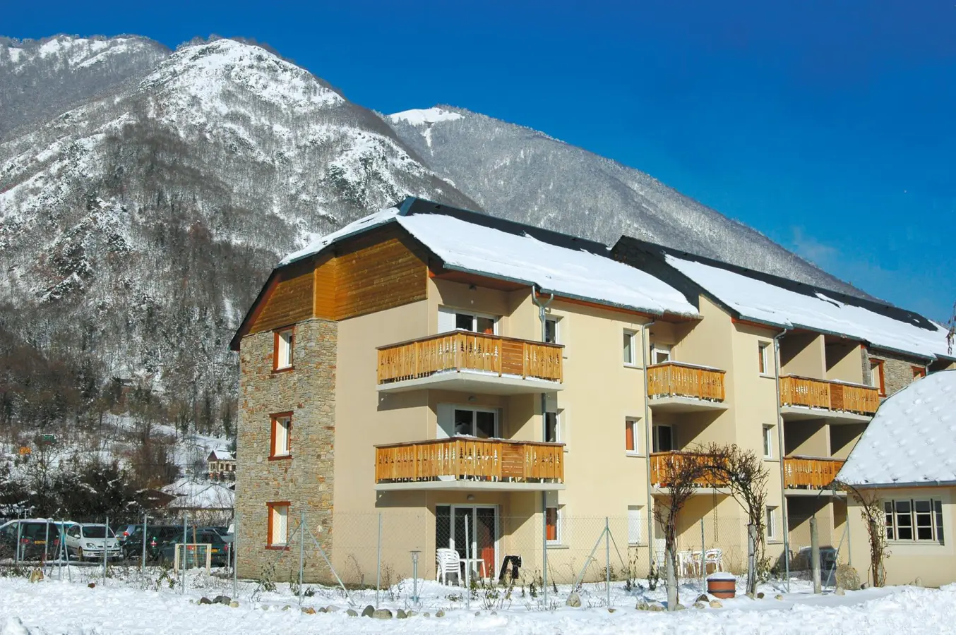Façade de la résidence Les Pics d’Aran, entourée de montagnes enneigées, avec balcons en bois et ciel bleu en arrière-plan