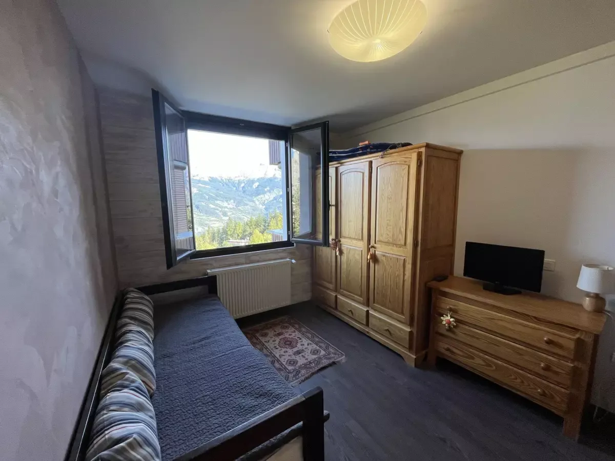 Chambre cosy avec lit simple, meubles en bois et fenêtre ouverte offrant une vue imprenable sur les montagnes alpines.