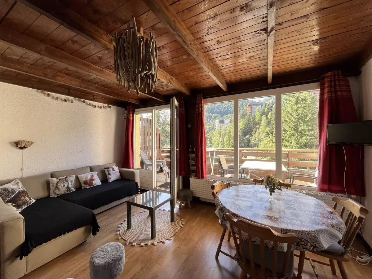 Salon chaleureux au plafond en bois avec canapé, table à manger et grande baie vitrée ouvrant sur un balcon avec vue sur la forêt alpine.