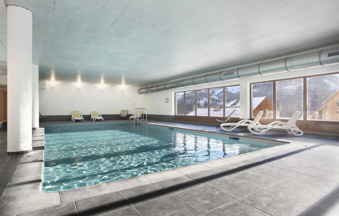 Piscine intérieure chauffée avec transats, offrant une vue panoramique sur les montagnes enneigées à travers de larges baies vitrées.