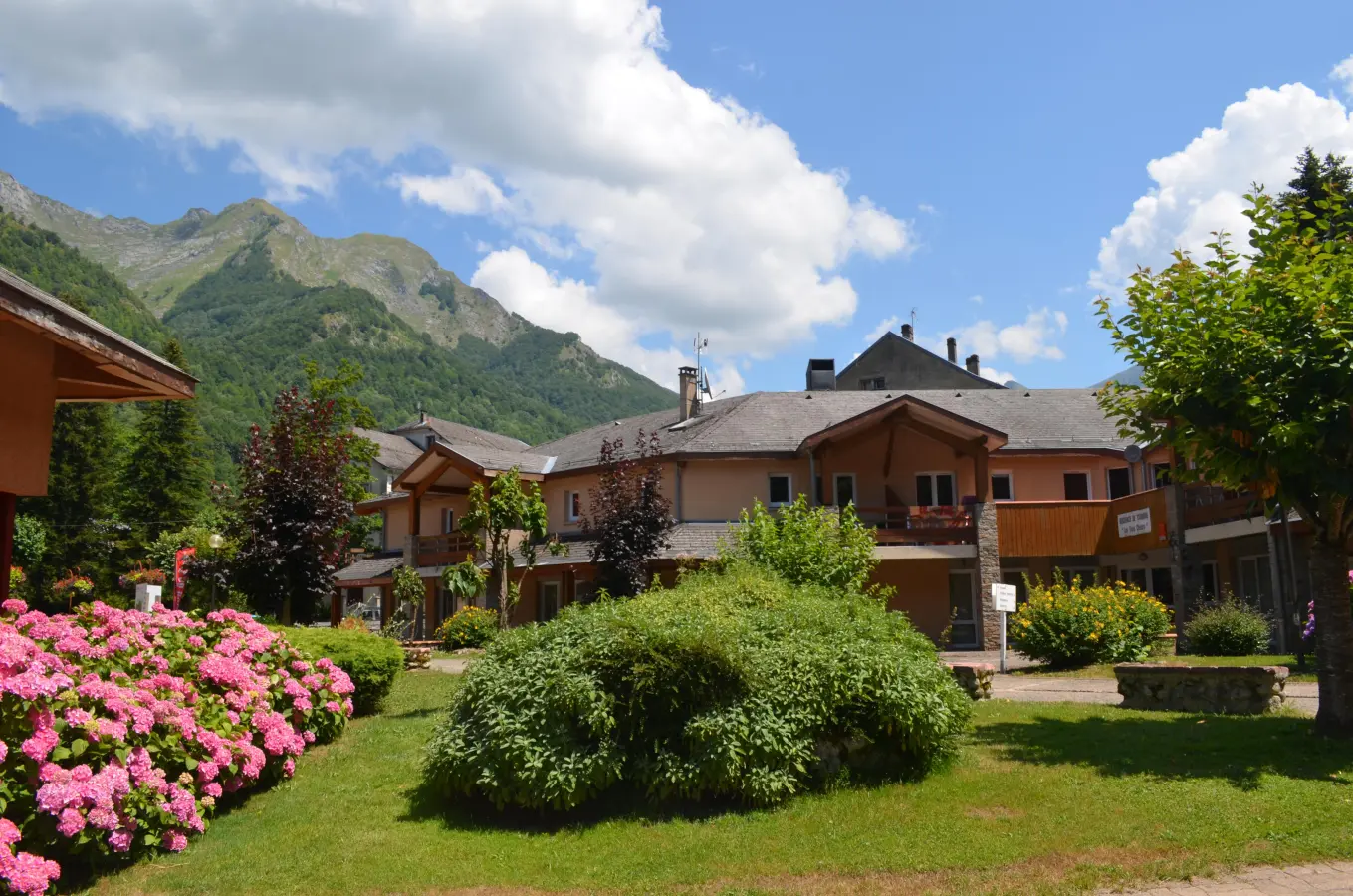 Résidence Les 3 Césars avec jardin fleuri, au pied des montagnes verdoyantes, sous un ciel bleu d'été.