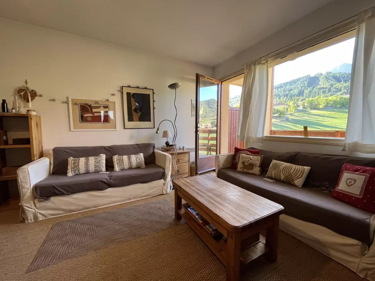 Salon chaleureux avec deux canapés, décoration montagnarde et grande fenêtre offrant une vue dégagée sur les paysages verdoyants alpins.