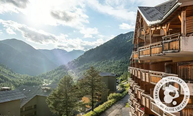 Vue panoramique depuis une résidence de montagne en bois surplombant une vallée verdoyante entourée de sommets alpins.