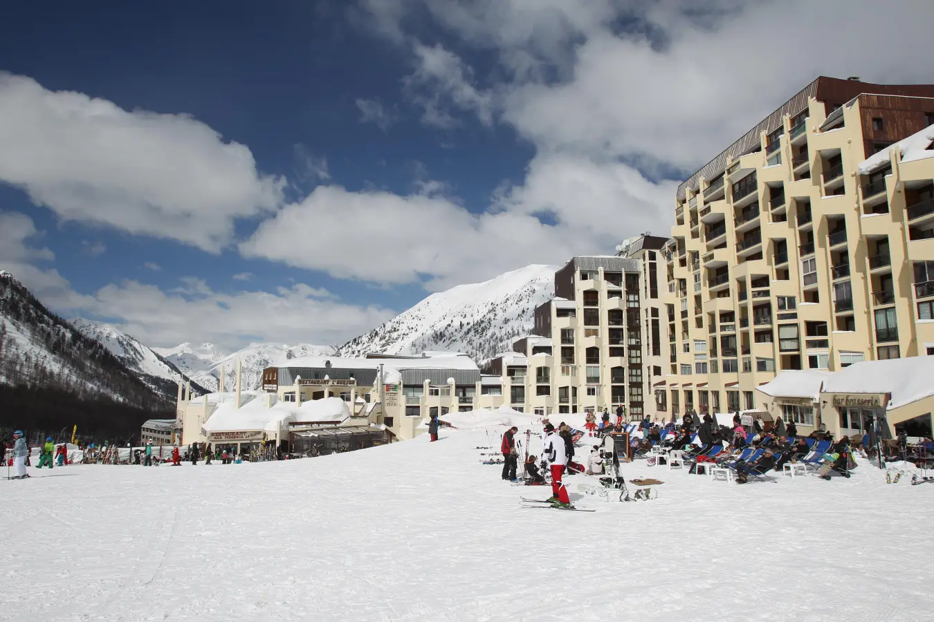 Résidence au pied des pistes enneigées avec skieurs et terrasses animées face aux montagnes des Alpes du Sud.