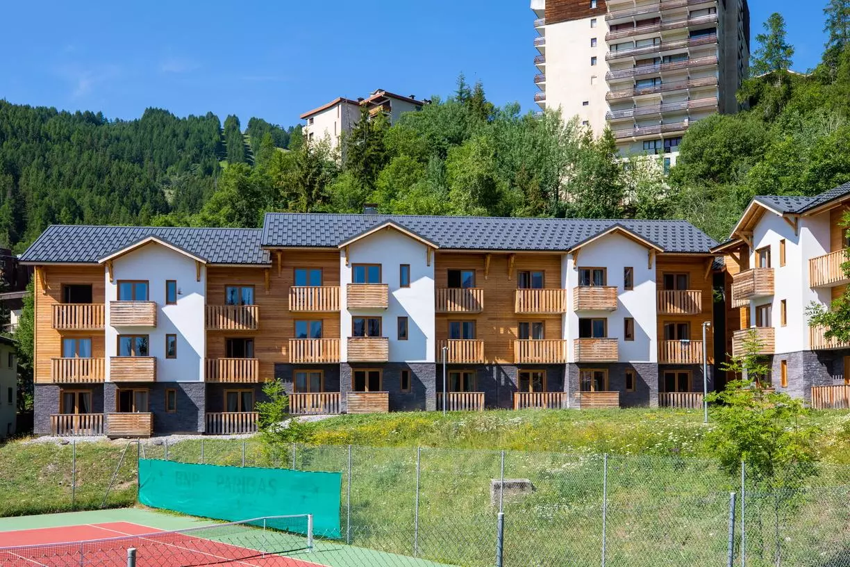 Façade moderne de la résidence Sowell à Pra Loup avec balcons en bois, entourée de verdure et située à proximité d’un court de tennis.